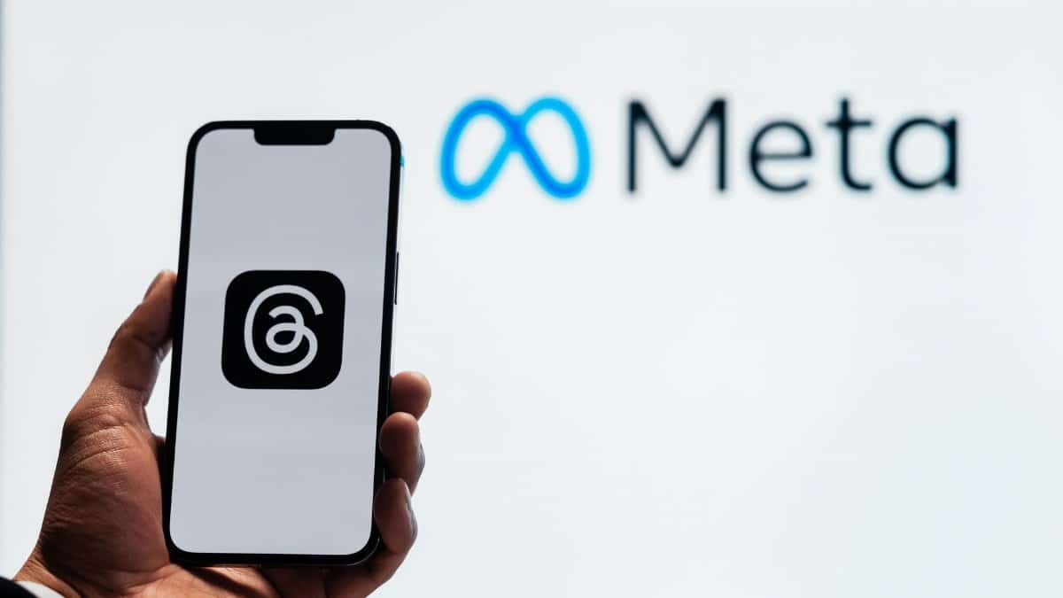 A hand holding a smartphone showing the Threads app with Meta logo in the background.