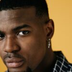 Close-up portrait of a serious man with earring against a vibrant yellow backdrop.