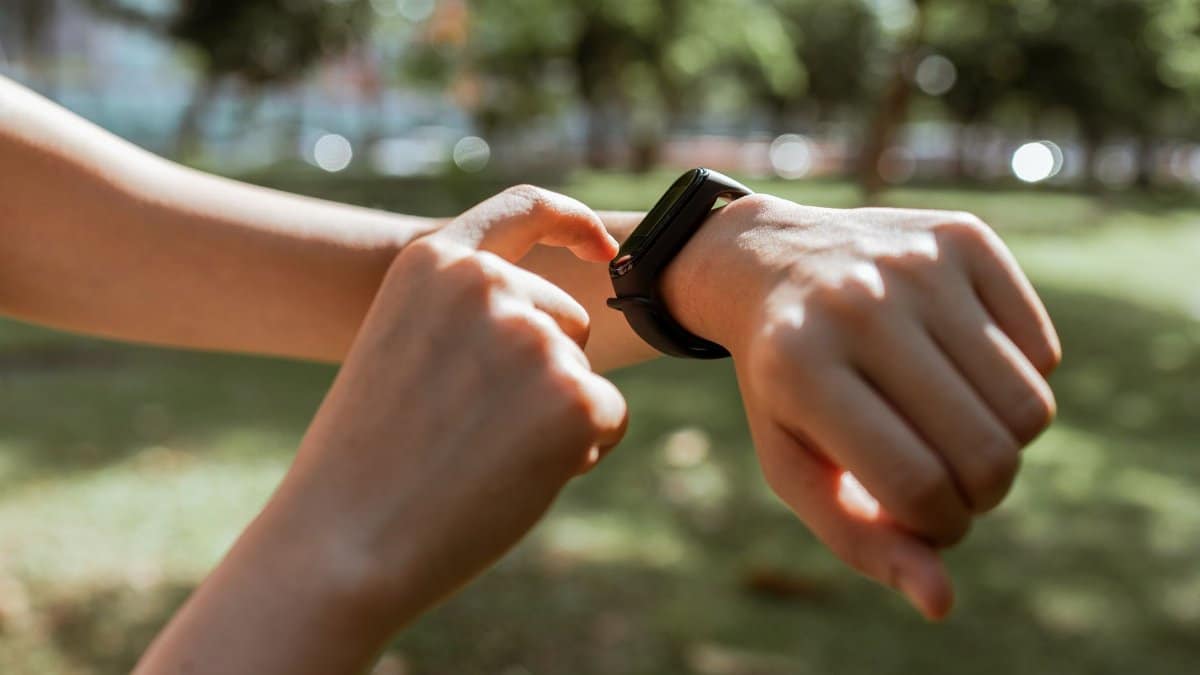 Crop faceless slim female tapping on fitness bracelet screen while training time in sunny summer park