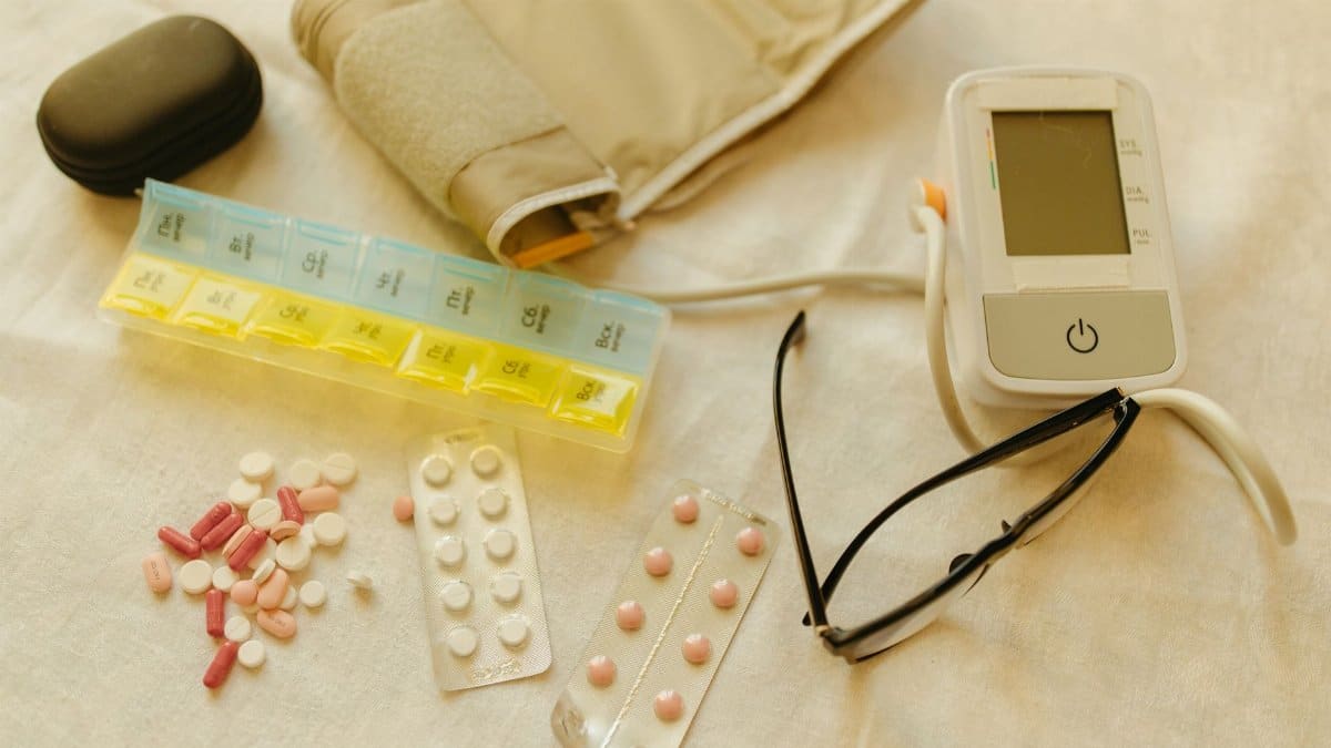 Flat lay image of healthcare essentials including a blood pressure monitor, pills, and eyeglasses on a white surface.