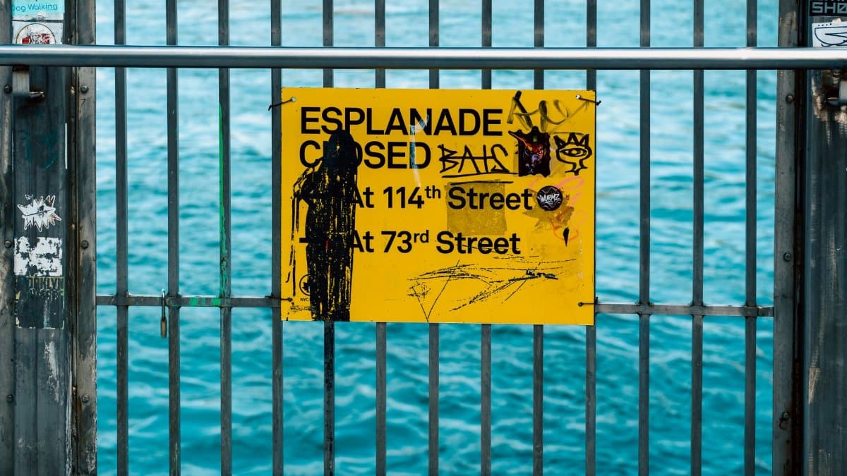 Yellow esplanade closure sign with graffiti on a metal railing by the waterfront in New York City.