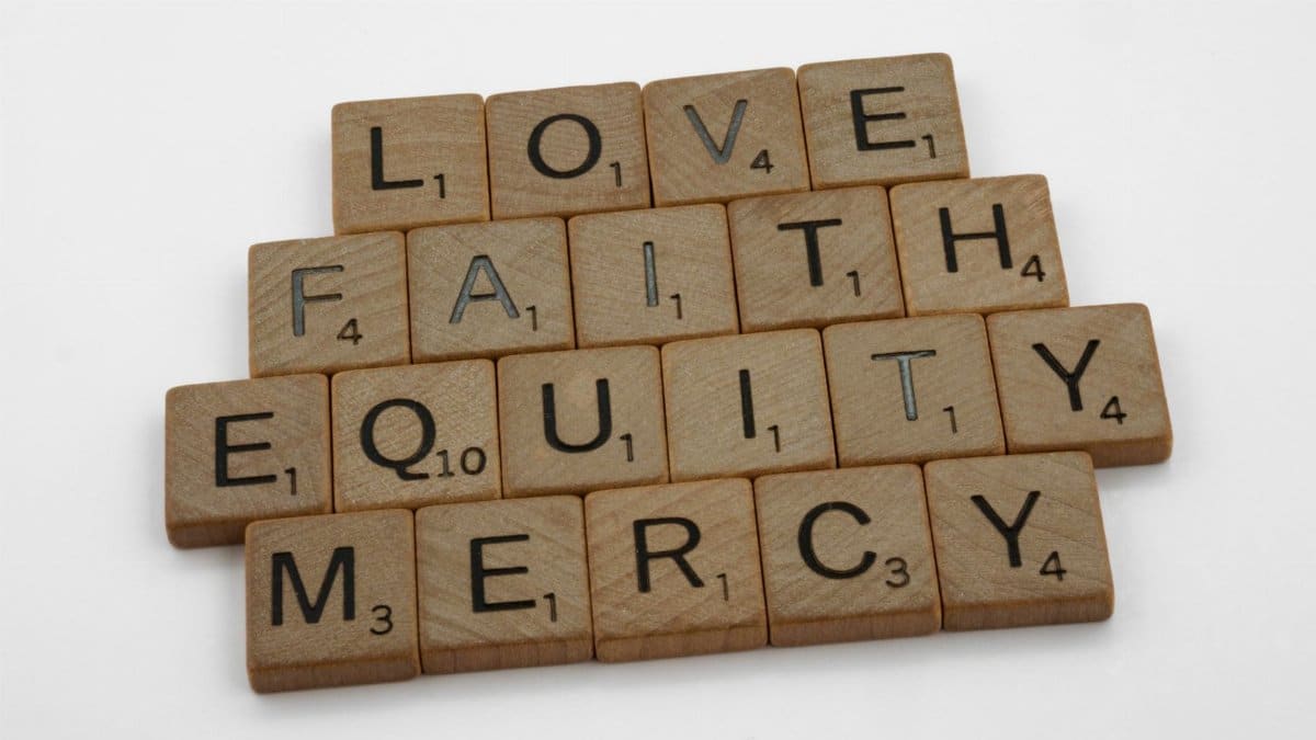 Wooden Scrabble tiles arranged to spell 'Love', 'Faith', 'Mercy', 'Equity' on a white background.