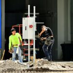 Two construction workers install an electrical panel outside a building site, wearing safety gear.