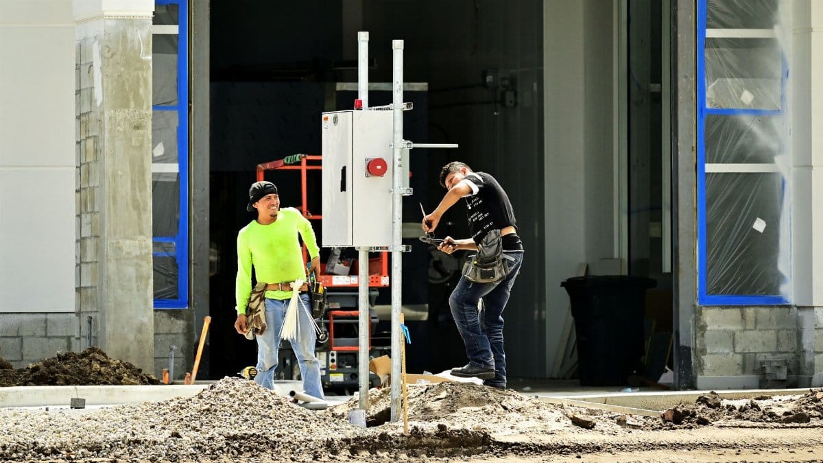 Two construction workers install an electrical panel outside a building site, wearing safety gear.