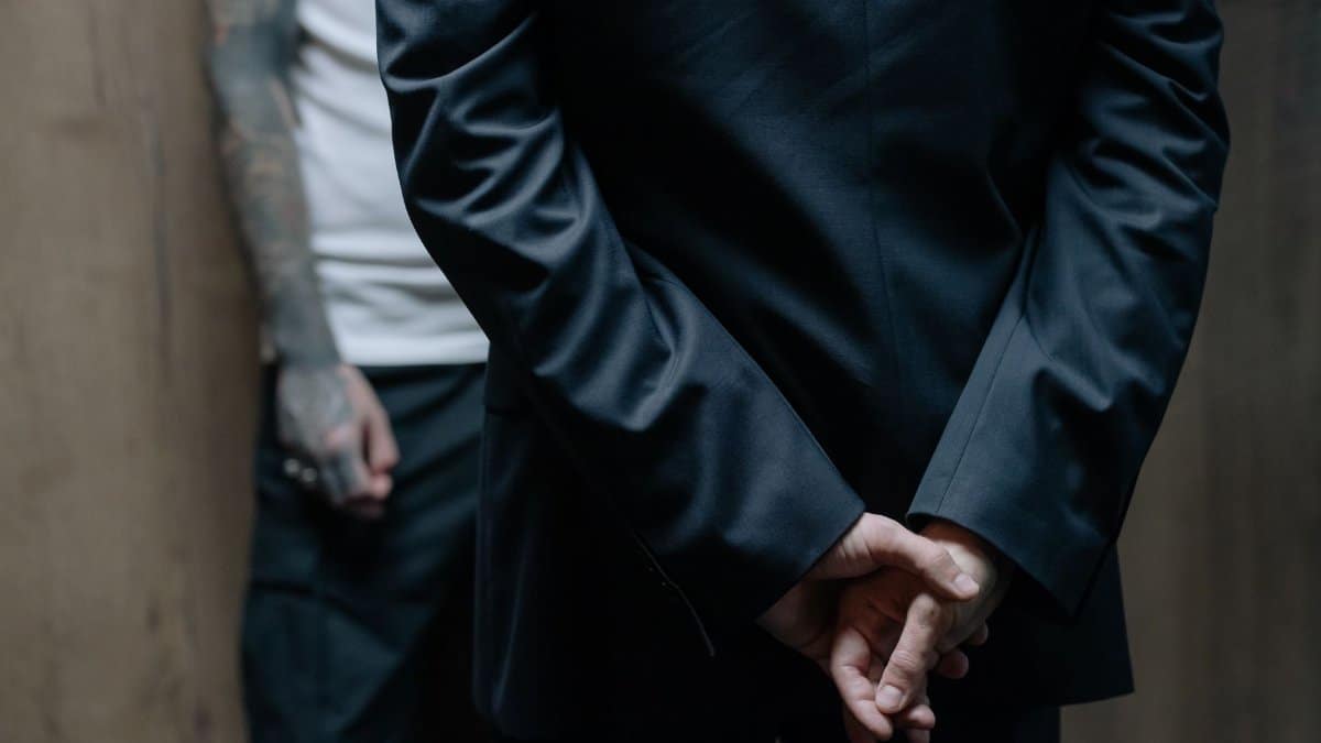 Close-up of man in suit with hands behind back and tattooed arm in view.