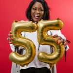 Smiling woman celebrating her 55th birthday with gold balloons against a red background.