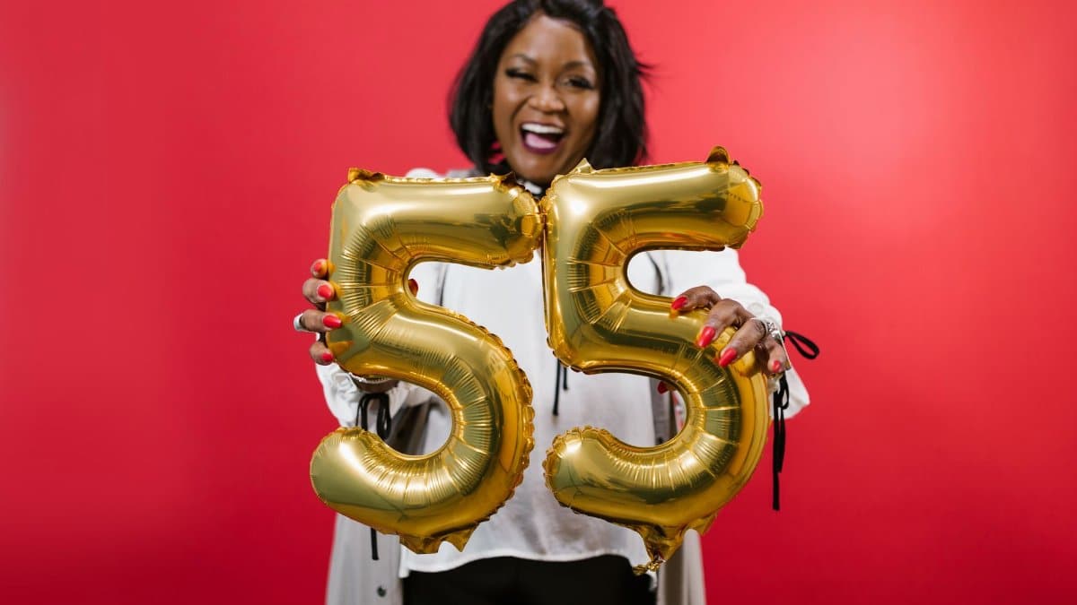 Smiling woman celebrating her 55th birthday with gold balloons against a red background.