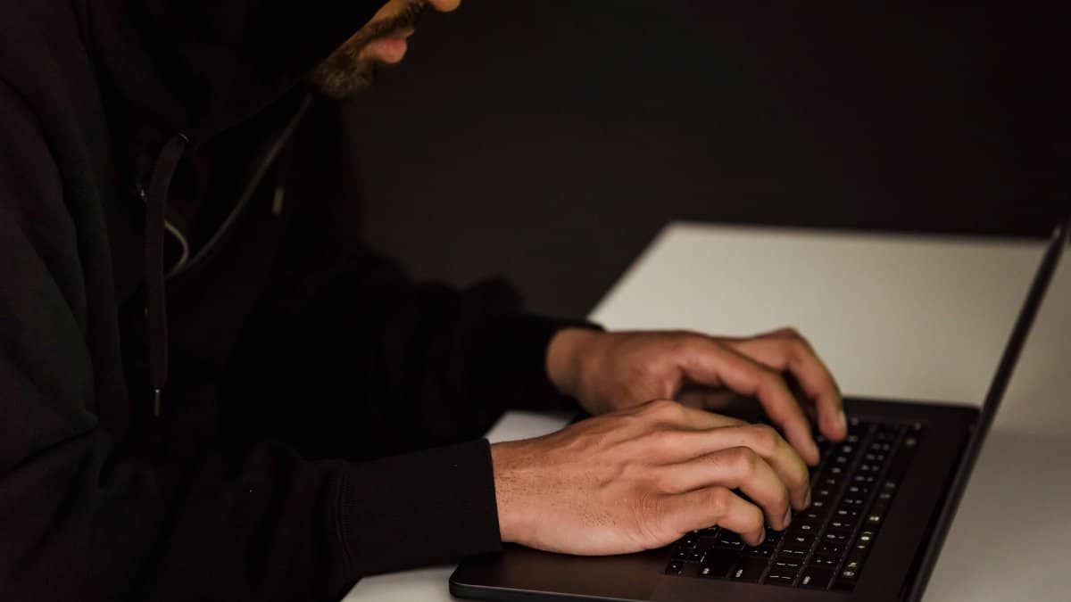 Crop bearded male hacker in black hood typing on netbook keyboard and browsing dark net in obscure room