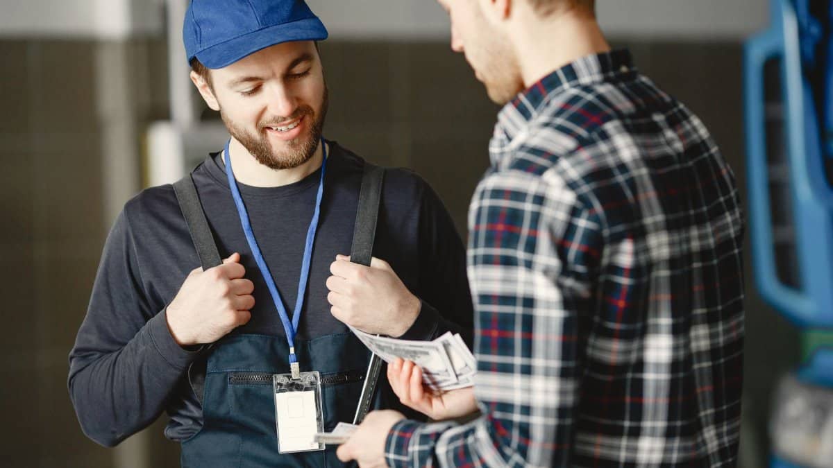 A mechanic in a blue cap receives cash payment from a customer in a plaid shirt, smiling.