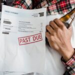 A man relaxes with a beer bottle while holding past due bills, highlighting financial stress.