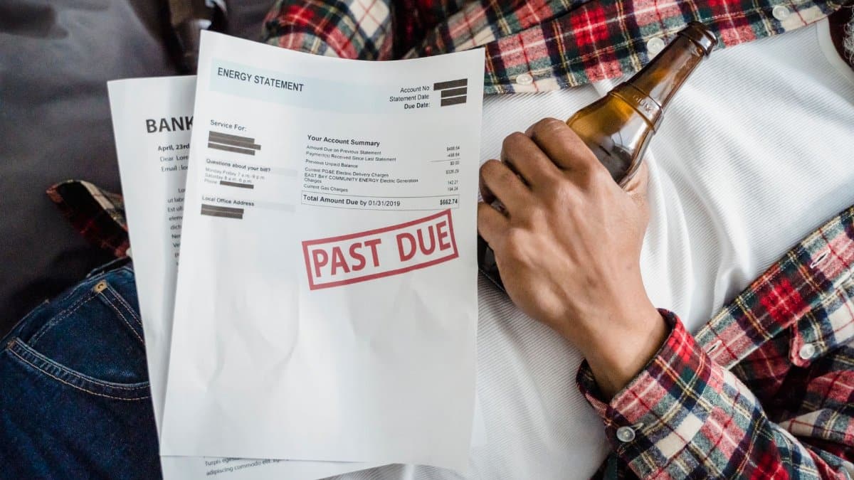 A man relaxes with a beer bottle while holding past due bills, highlighting financial stress.