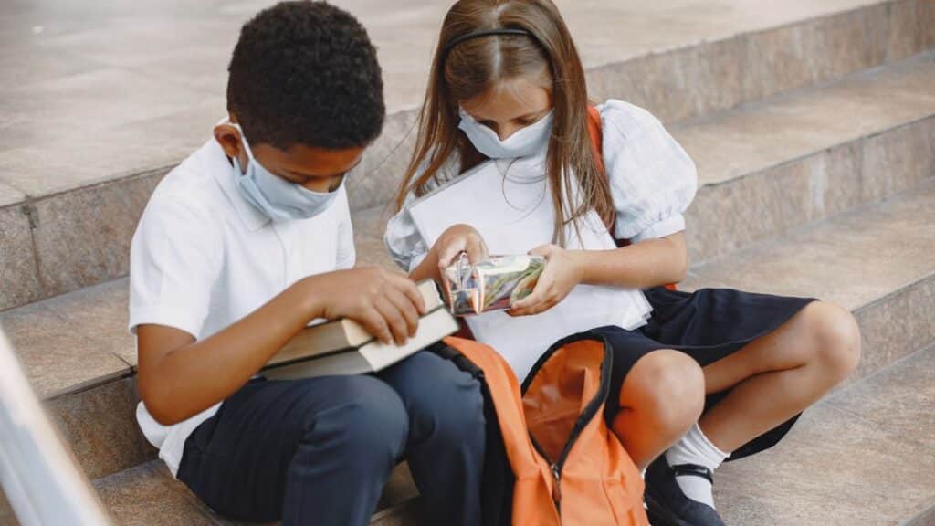 Two children wearing face masks and school uniforms, sitting on stairs with backpacks.