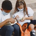 Two children wearing face masks and school uniforms, sitting on stairs with backpacks.