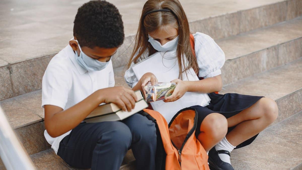 Two children wearing face masks and school uniforms, sitting on stairs with backpacks.