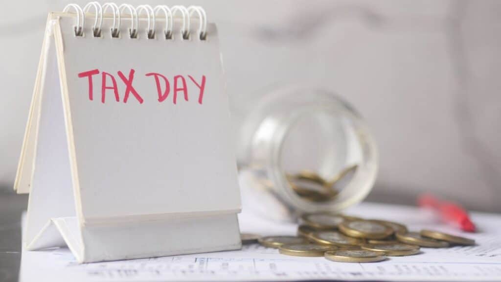 A close-up of a table calendar marked 'Tax Day' with a jar of coins and notes, symbolizing financial planning.