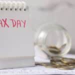 A close-up of a table calendar marked 'Tax Day' with a jar of coins and notes, symbolizing financial planning.
