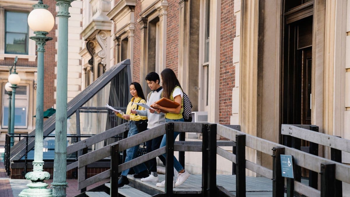 Three students walk down steps outside a university building, holding books and papers.