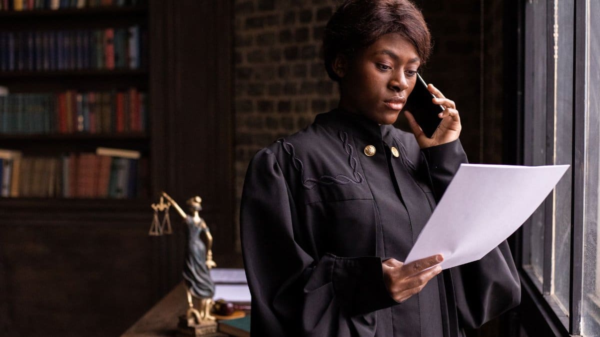 African American judge in robe, reading document and talking on phone by the office window.