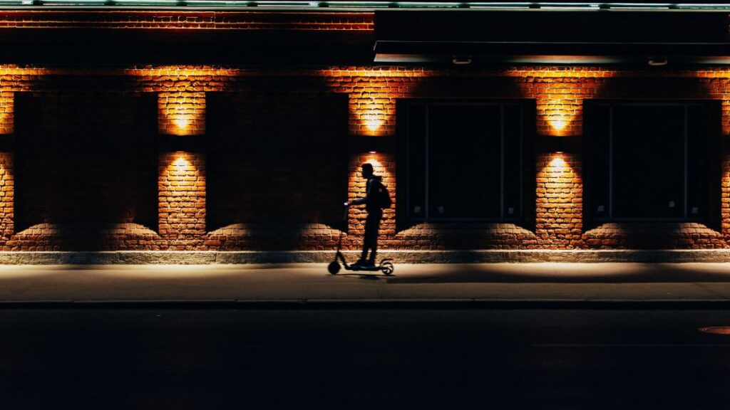 A silhouette of a person riding a scooter against a brick wall illuminated at night.