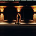 A silhouette of a person riding a scooter against a brick wall illuminated at night.
