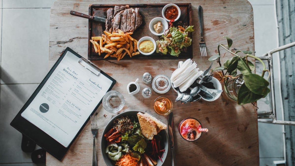 Top view of wooden table with salad bowl and fresh drink arranged with tray of appetizing steak and french fries near menu in cozy cafe