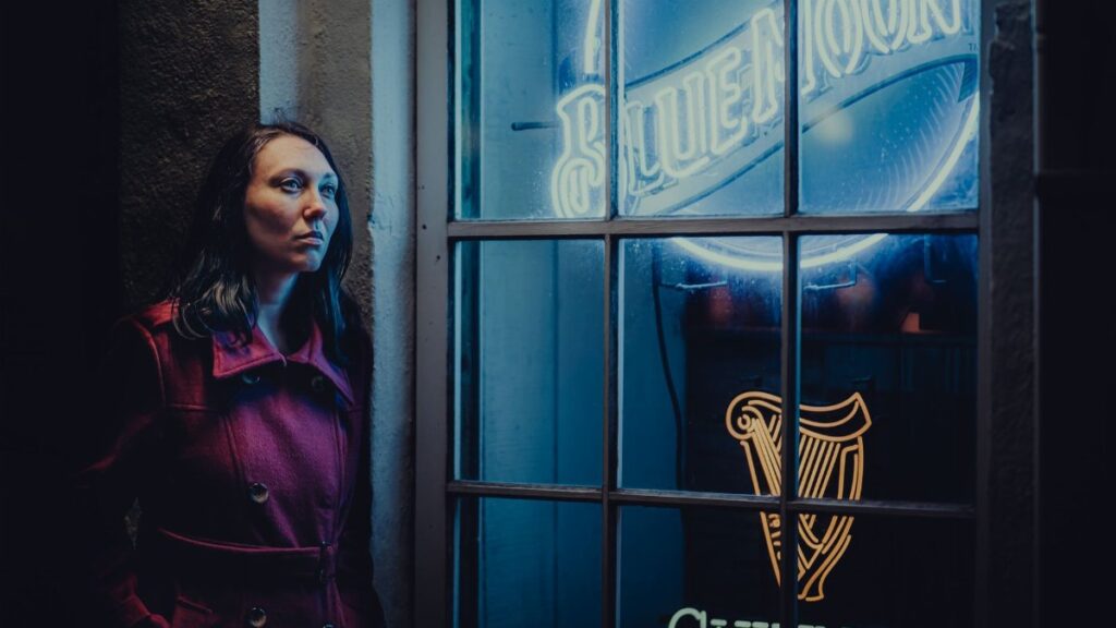 Woman in a red coat leans against a wall by blue neon signs at night.