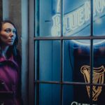 Woman in a red coat leans against a wall by blue neon signs at night.