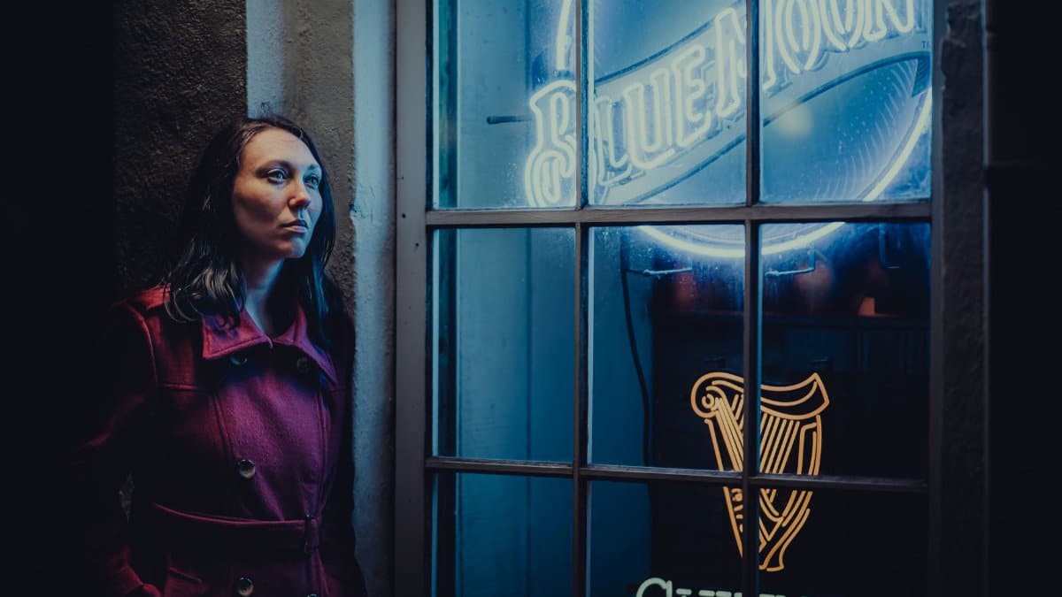 Woman in a red coat leans against a wall by blue neon signs at night.