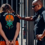 Police officer in sunglasses detains handcuffed individual in orange shorts outdoors.