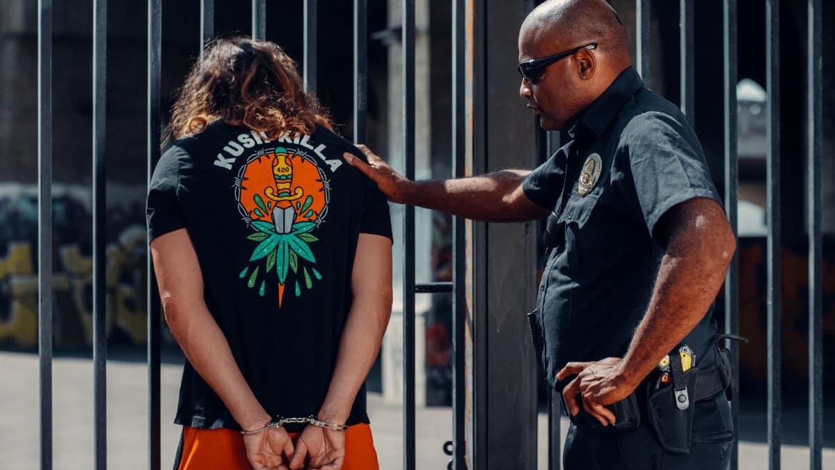 Police officer in sunglasses detains handcuffed individual in orange shorts outdoors.