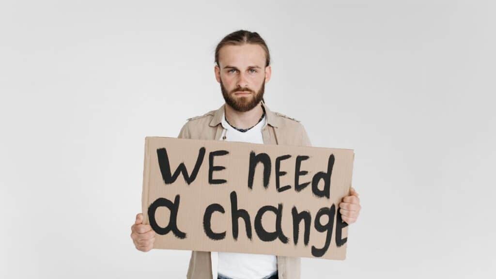 A man holding a protest sign reading 'We Need a Change' on a white background.