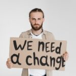 A man holding a protest sign reading 'We Need a Change' on a white background.