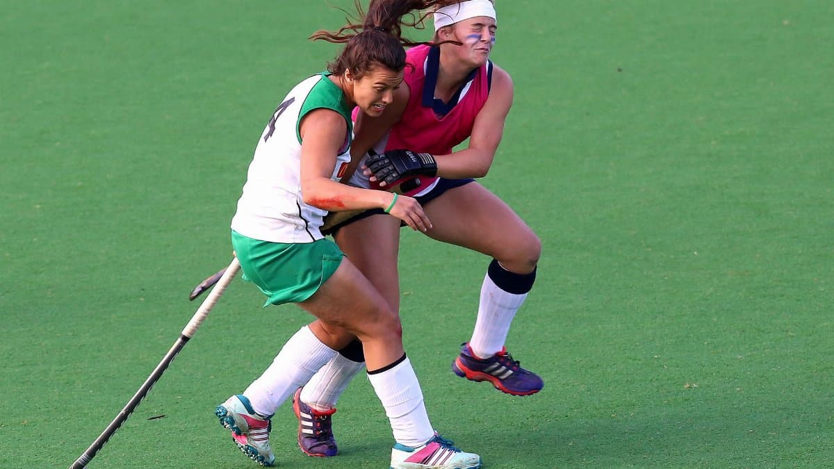 Two female field hockey players engage fiercely during a match on grass.