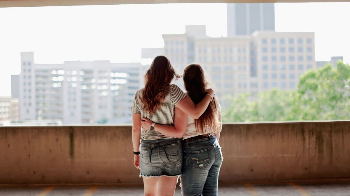 Two women in casual clothing embrace with a city skyline in the background.
