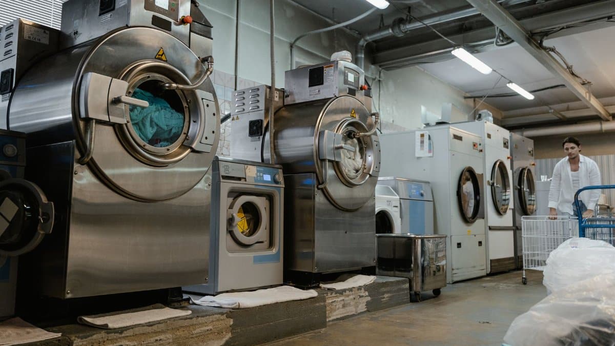Industrial laundromat interior with large washing machines and a man pushing a trolley.