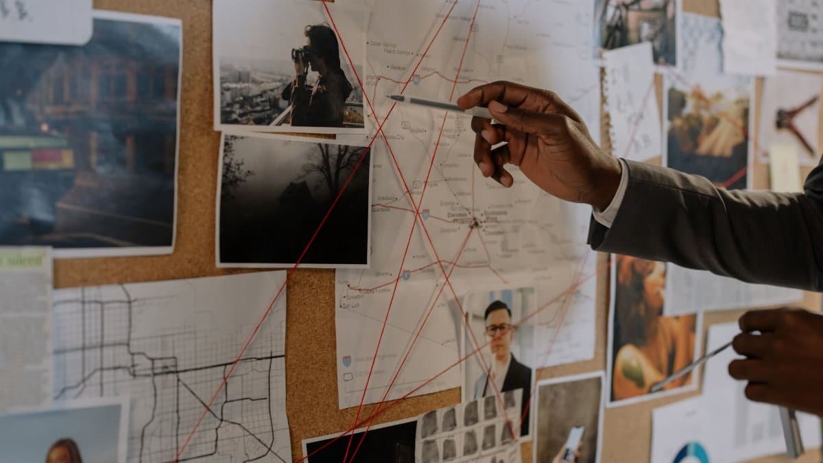 Detective examines a corkboard with maps and photos to solve a mystery.