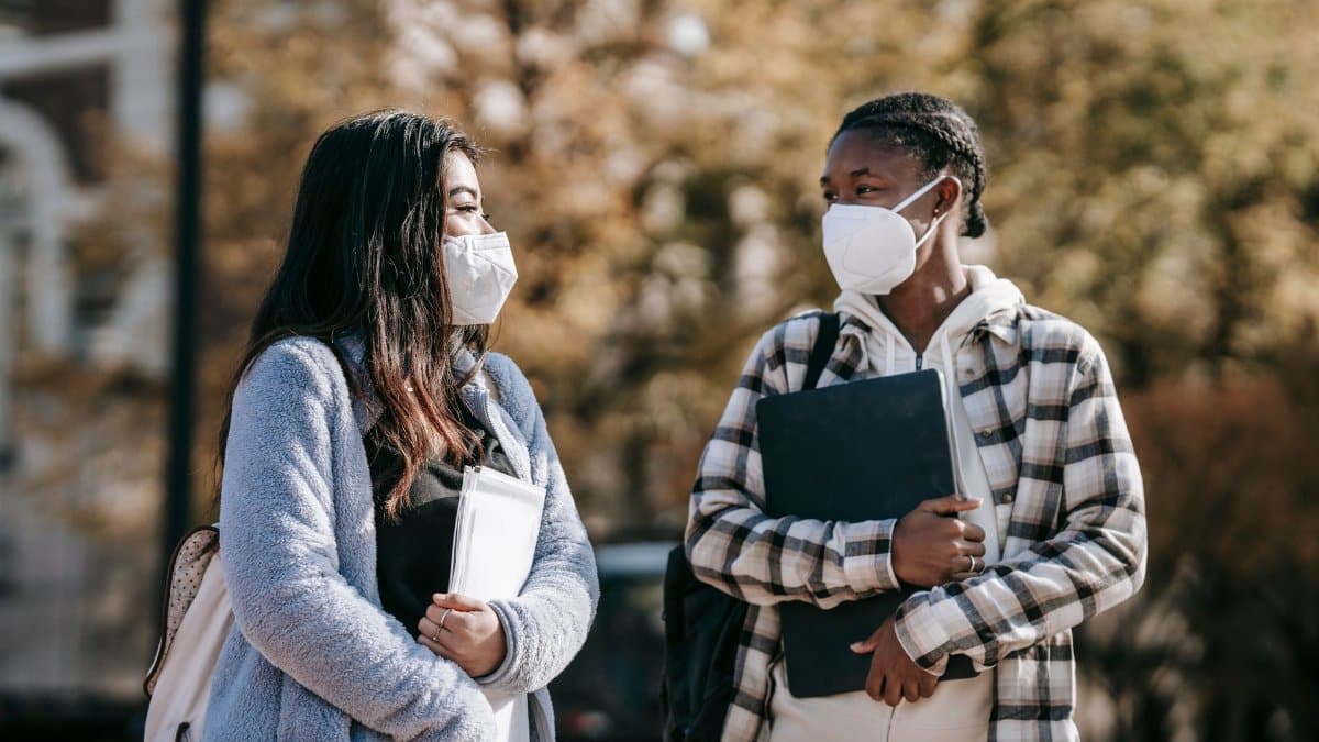 Young multiracial female students in casual outfit and protective masks with documents and laptop walking in university campus
