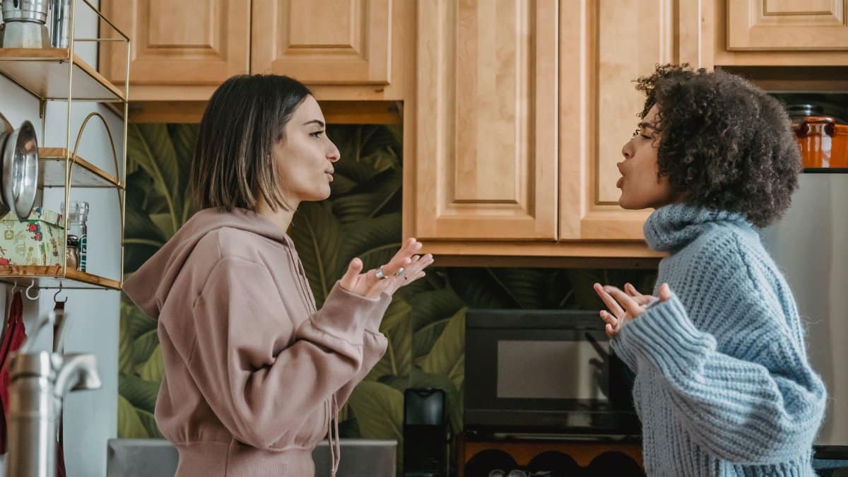 Side view of unhappy young multiracial girlfriends in casual clothes standing in kitchen while having disagreement and looking at each other