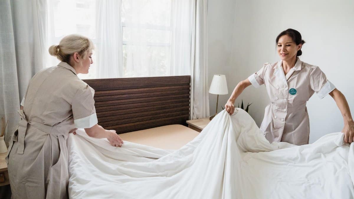 Two hotel housekeepers changing bed sheets in a sunlit room. Professional cleaning and hospitality.