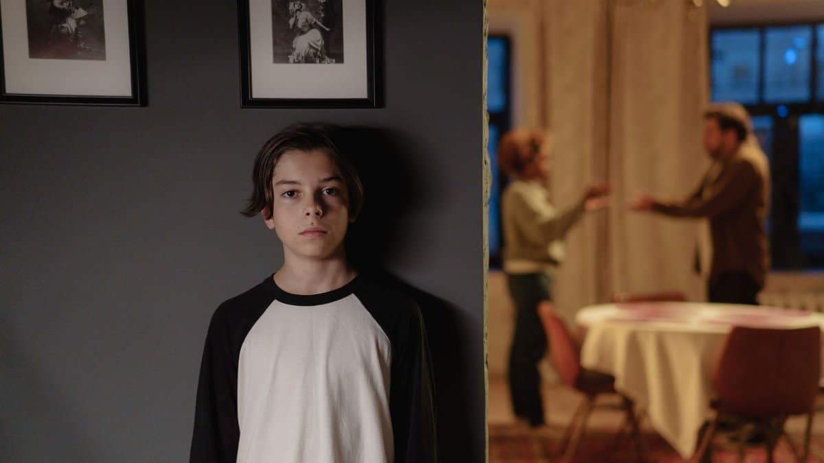 A boy stands in focus, while two adults argue in the background, depicting family conflict.