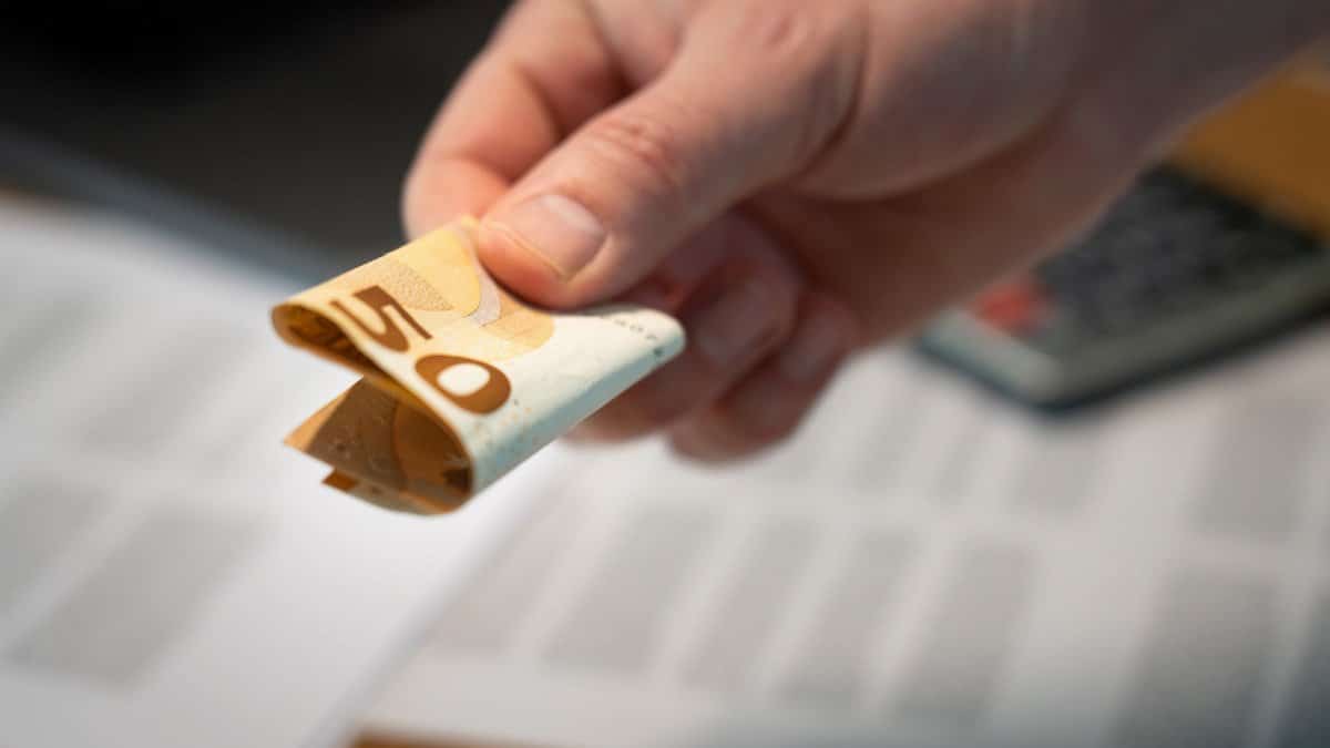 A detailed image of a hand holding a folded 50 euro banknote, with documents in the background.