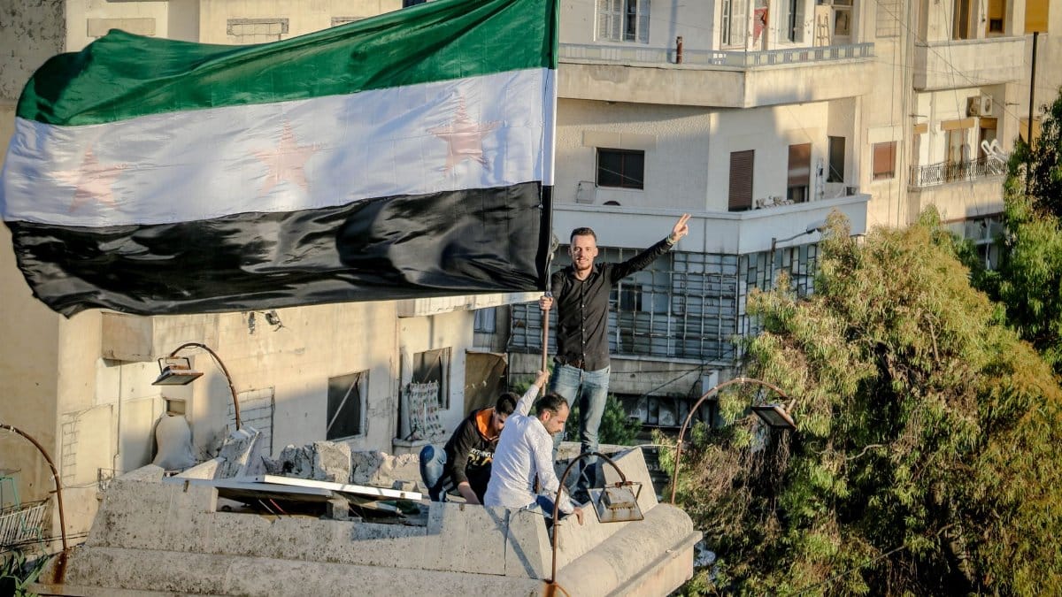 Arab man with national flag demonstrating victory gesture against friends on building rooftop in town