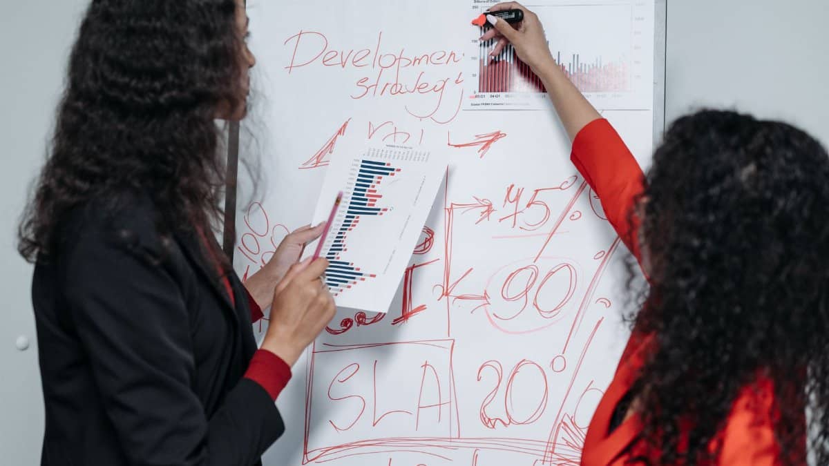 Two professional women discuss and strategize on business development in a modern office setting.