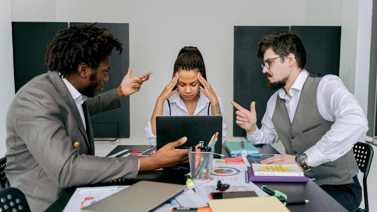 Two men argue while a woman looks frustrated at a laptop in an office environment.