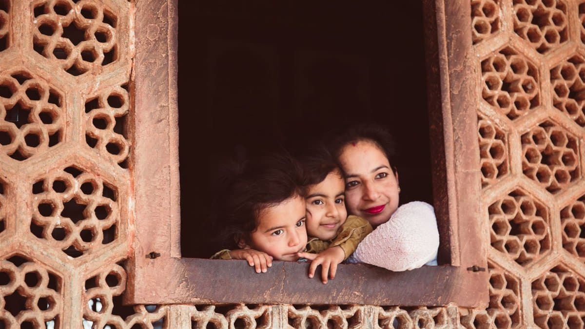 A mother and her two children gaze outside from an intricately designed stone window, embracing their togetherness.
