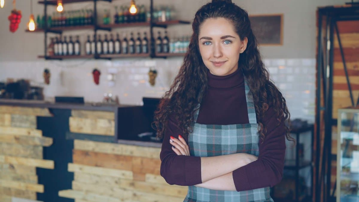Confident café owner stands with arms crossed inside a rustic, cozy coffee shop.