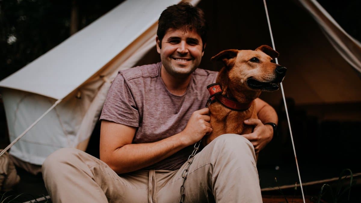 A man sitting joyfully with his dog outside a tent in a camping setting.
