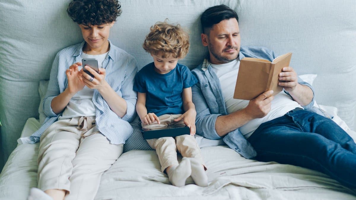 A family relaxing on bed with a smartphone, tablet, and book, symbolizing modern family life.