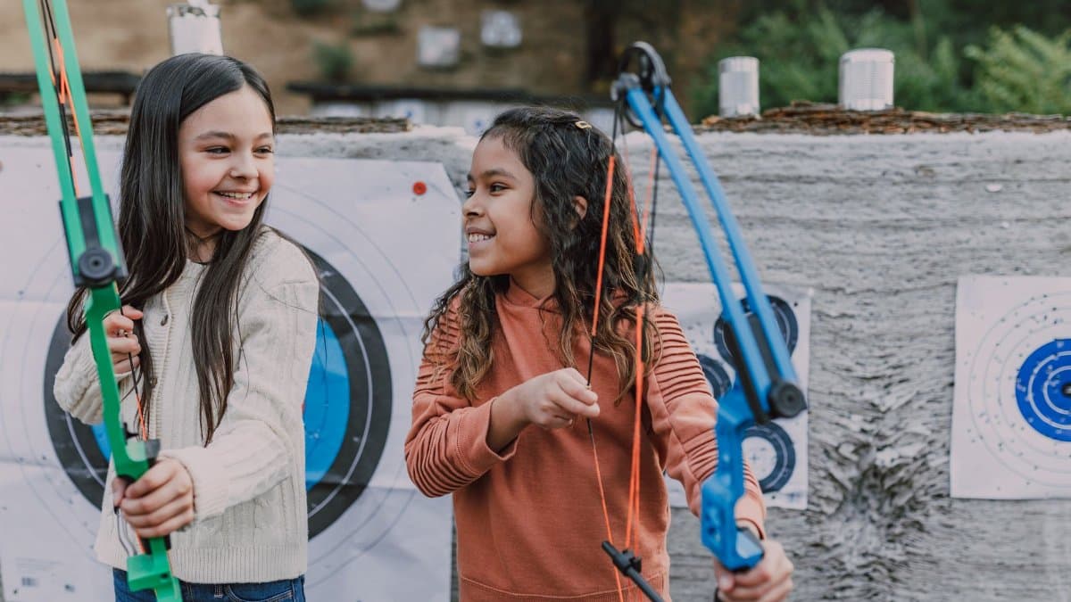 Two girls enjoying archery practice outdoors with colorful bows and target boards.