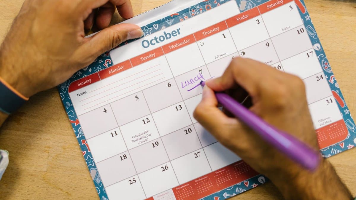 Close-up of a person writing a lunch reminder on an October calendar with a purple pen.